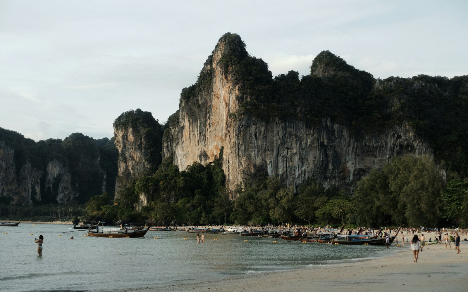 Longtail boats and families at a beach beneath limestone cliffs in Krabi, Thailand
